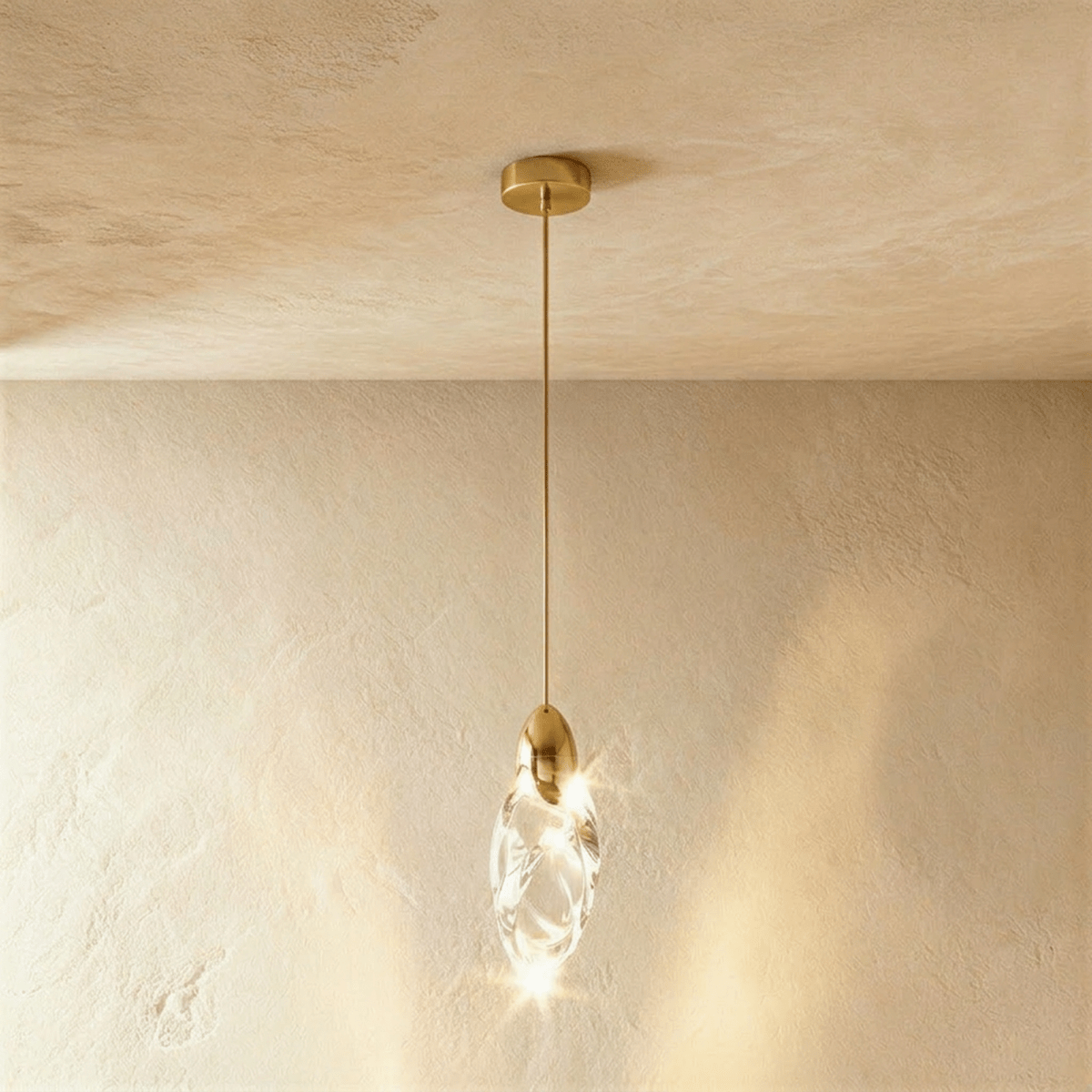 Clérachic lit clear glass teardrop pendant with a polished brass cap and slim brass stem, glowing against a plaster wall.
