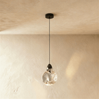 Clérachic round rippled clear glass pendant with a dark metal cap on a black cord and canopy, against a warm plaster wall.