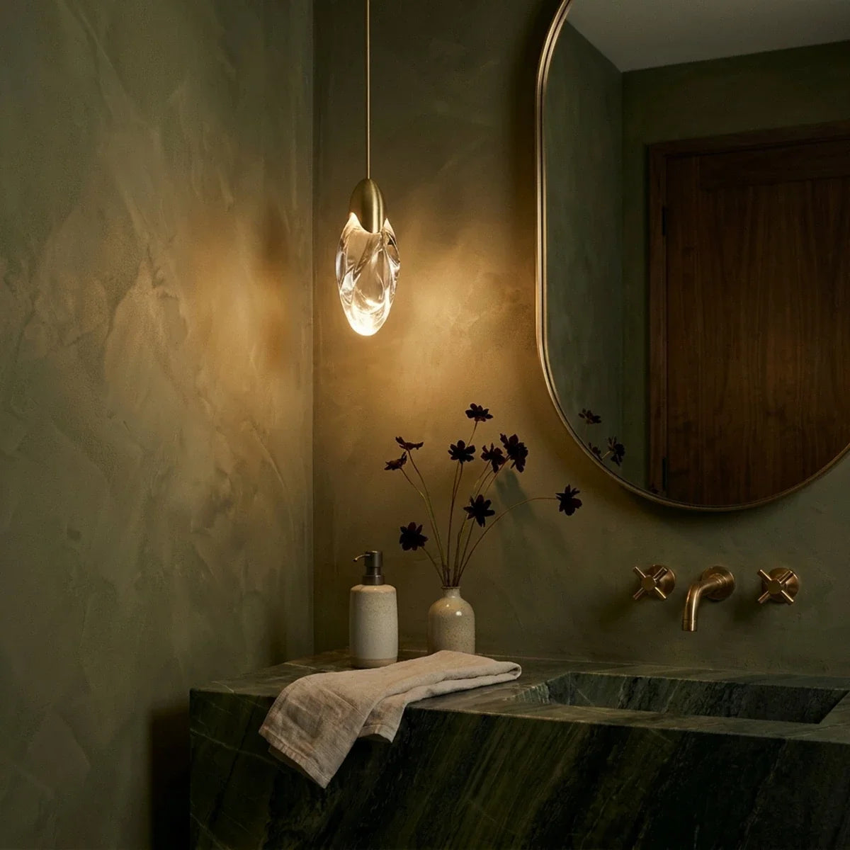Clérachic brass-capped glass teardrop pendant glowing beside an oval mirror above a green marble washstand.