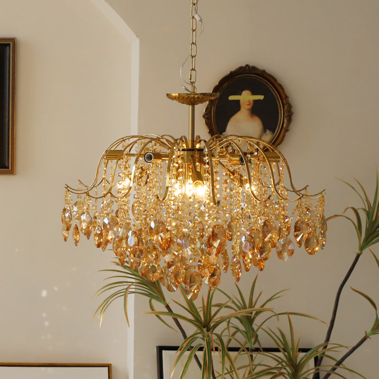 Amber crystals glow on a Clérachic chandelier hung from a chain, with a framed portrait and a tall green plant behind it.