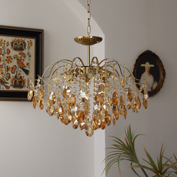 A Clérachic amber crystal chandelier on a chain, beside a folk-pattern framed textile and a round portrait in a white-walled corner.