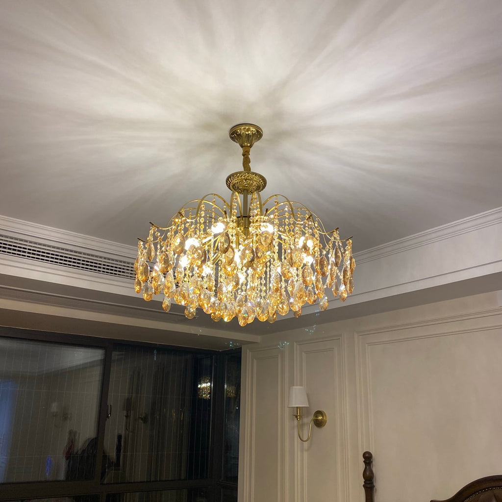 Gold chandelier from Clérachic lit under a white ceiling with a patterned light spread, above a room with a wall light and tall window.