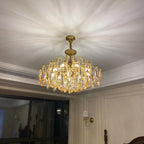 Gold chandelier from Clérachic lit under a white ceiling with a patterned light spread, above a room with a wall light and tall window.
