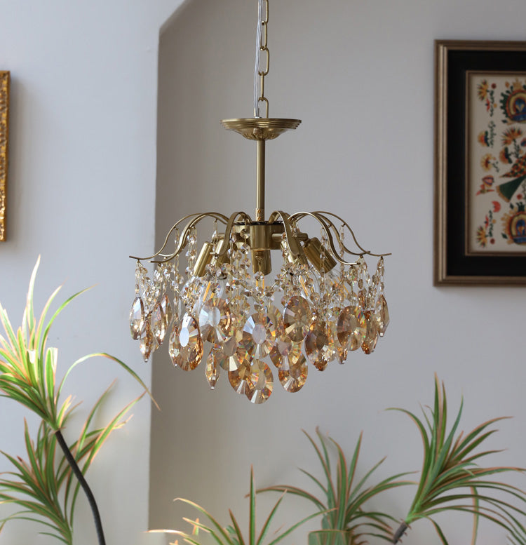 Chain-hung Clérachic chandelier with amber crystals, set against a white wall with a framed folk print and spiky plant leaves.