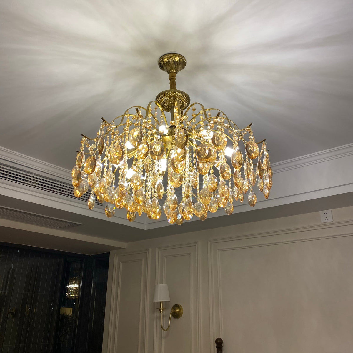 Clérachic chandelier switched on in a panelled room, throwing a soft starburst of light across the ceiling above a wall sconce.