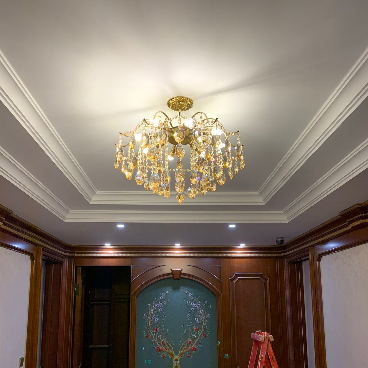 This Clérachic crystal chandelier lit in a recessed ceiling with white cornice, above a room with dark wood panelling and a teal arched door.