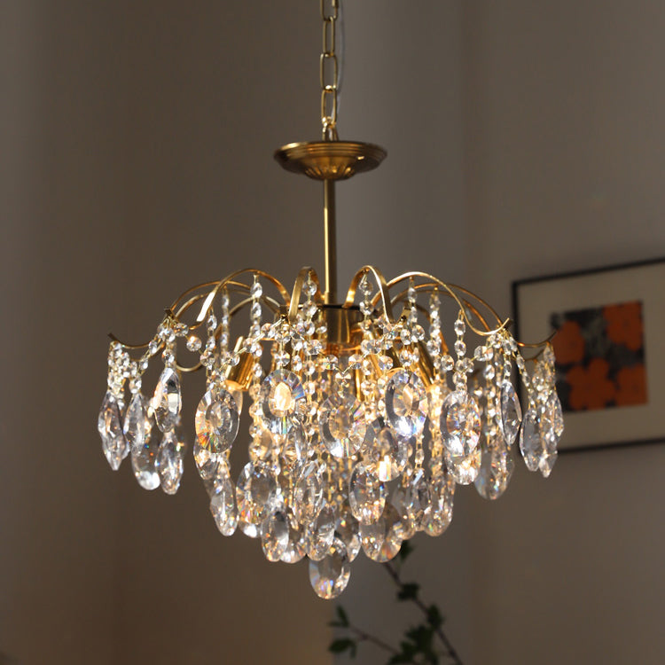 Clérachic gold chandelier with clear crystals and glowing bulbs, hung on a chain beside a framed print with orange tones.