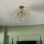 Ceiling-mounted Clérachic chandelier with clear teardrop crystals, seen against a white ceiling above a sage-green wall and a dark bed frame.