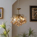 A Clérachic chandelier with amber crystals lit beside a framed folk-art textile, with green plant leaves in the foreground.