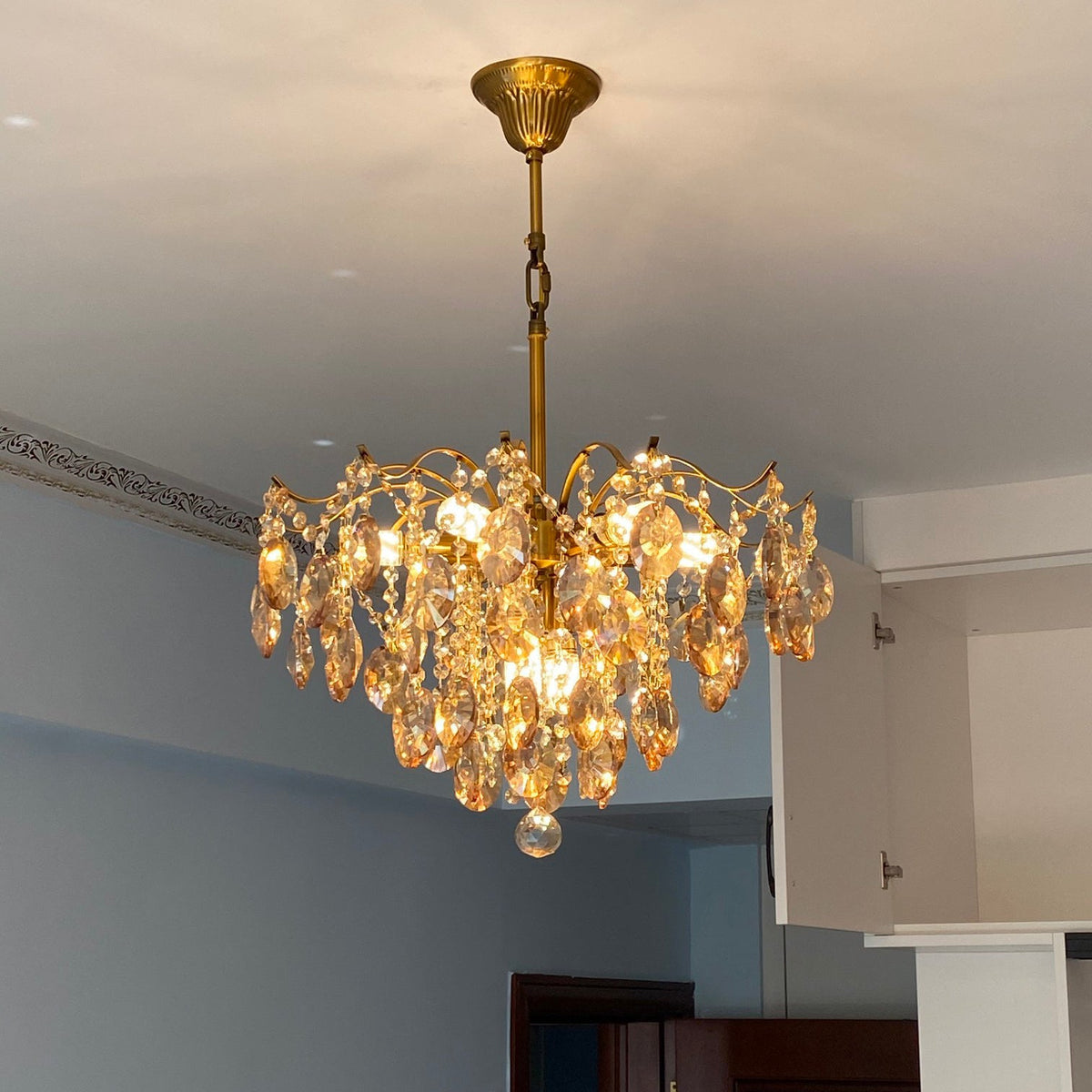 Clérachic chandelier with amber crystals hung on a chain below a white ceiling, beside a blue wall and a kitchen cabinet.