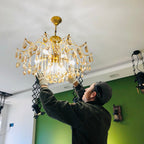 Someone in a dark cap and gloves lifts this Clérachic crystal chandelier into place under a white ceiling, with a green wall behind.