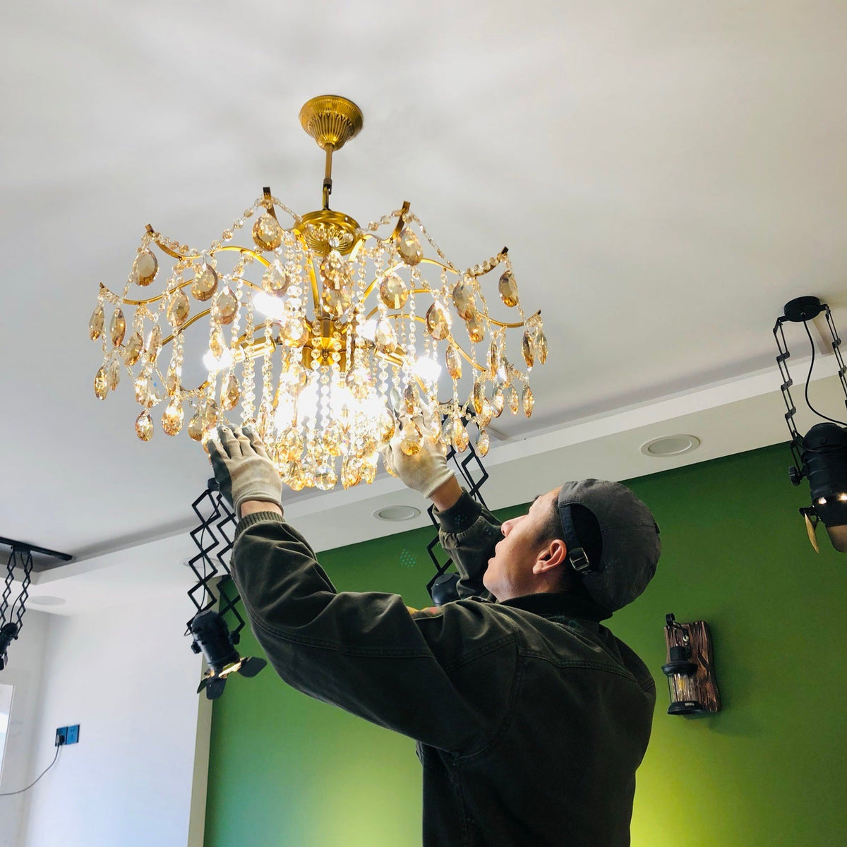 Someone in a dark cap and gloves lifts this Clérachic crystal chandelier into place under a white ceiling, with a green wall behind.