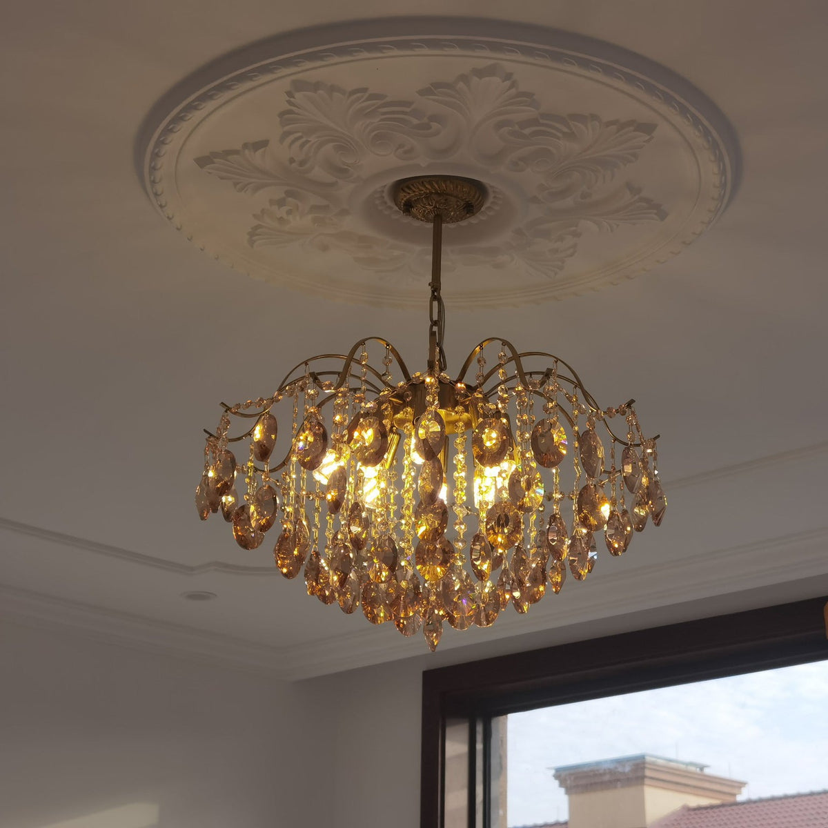 Amber crystal chandelier from Clérachic hung beneath a carved white ceiling rose, with a window to one side.