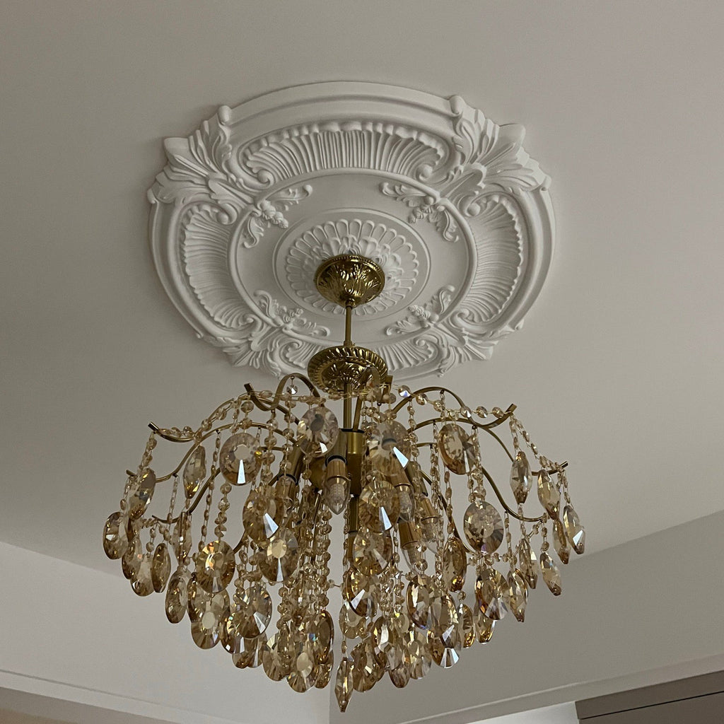 View from below of this Clérachic chandelier under an ornate white ceiling rose, its crystals and brass arms catching the light.