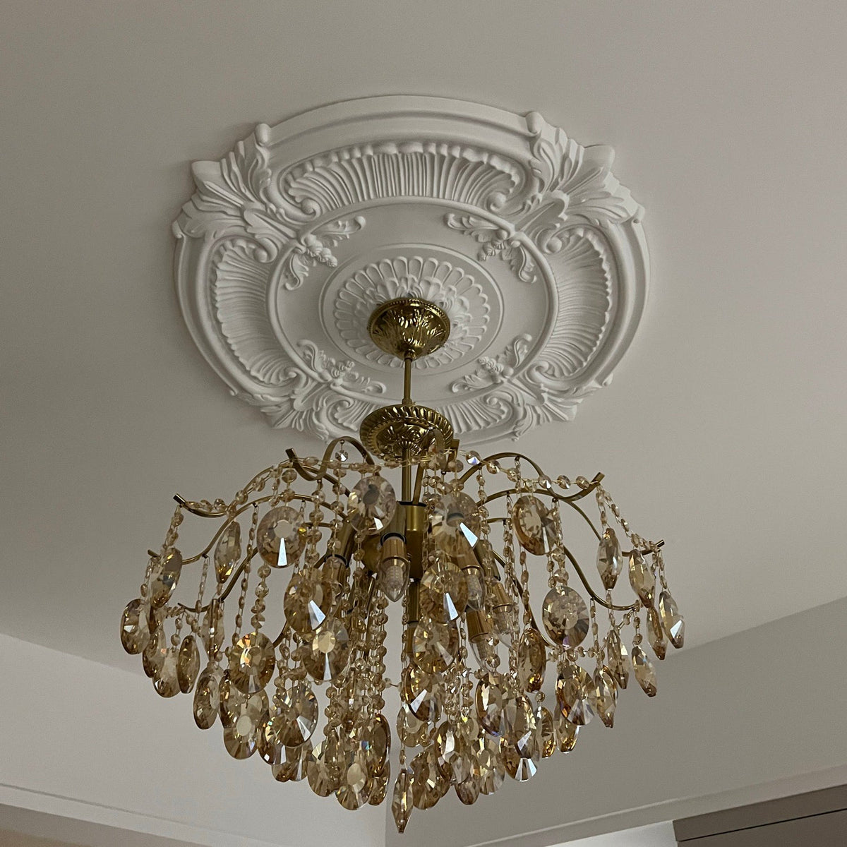 View from below of this Clérachic chandelier under an ornate white ceiling rose, its crystals and brass arms catching the light.