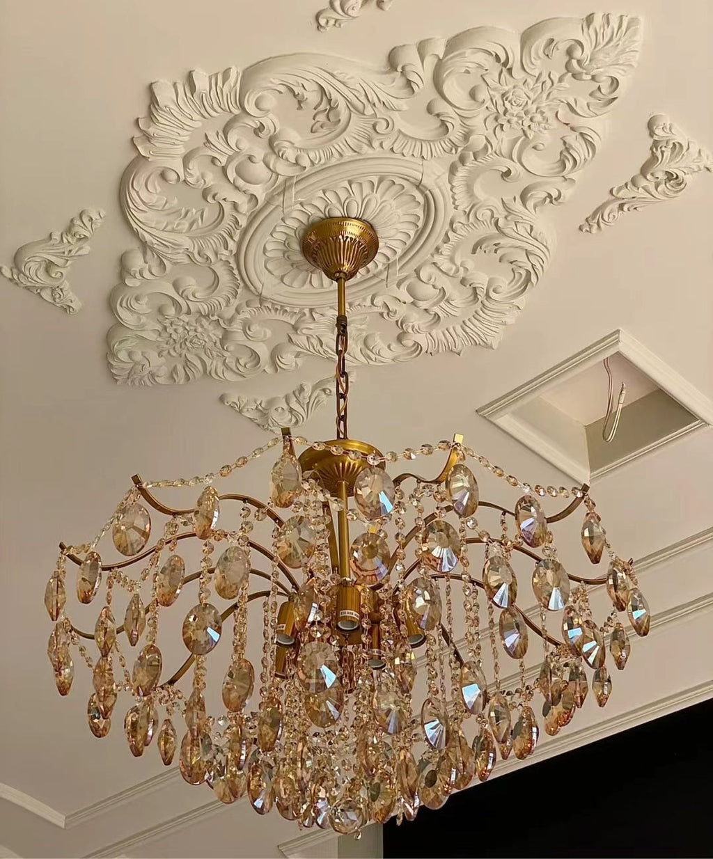 View from below of a Clérachic chandelier with amber crystals, under a detailed white plaster ceiling rose beside a recessed panel.