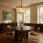 Clérachic tiered crystal-rod chandelier above a round dark dining table with tan leather chairs and tall sash windows.