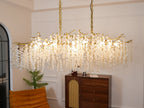Wide view of the lit Clérachic chandelier in a white-panelled room, above a walnut cabinet with a rattan door and an orange chair.