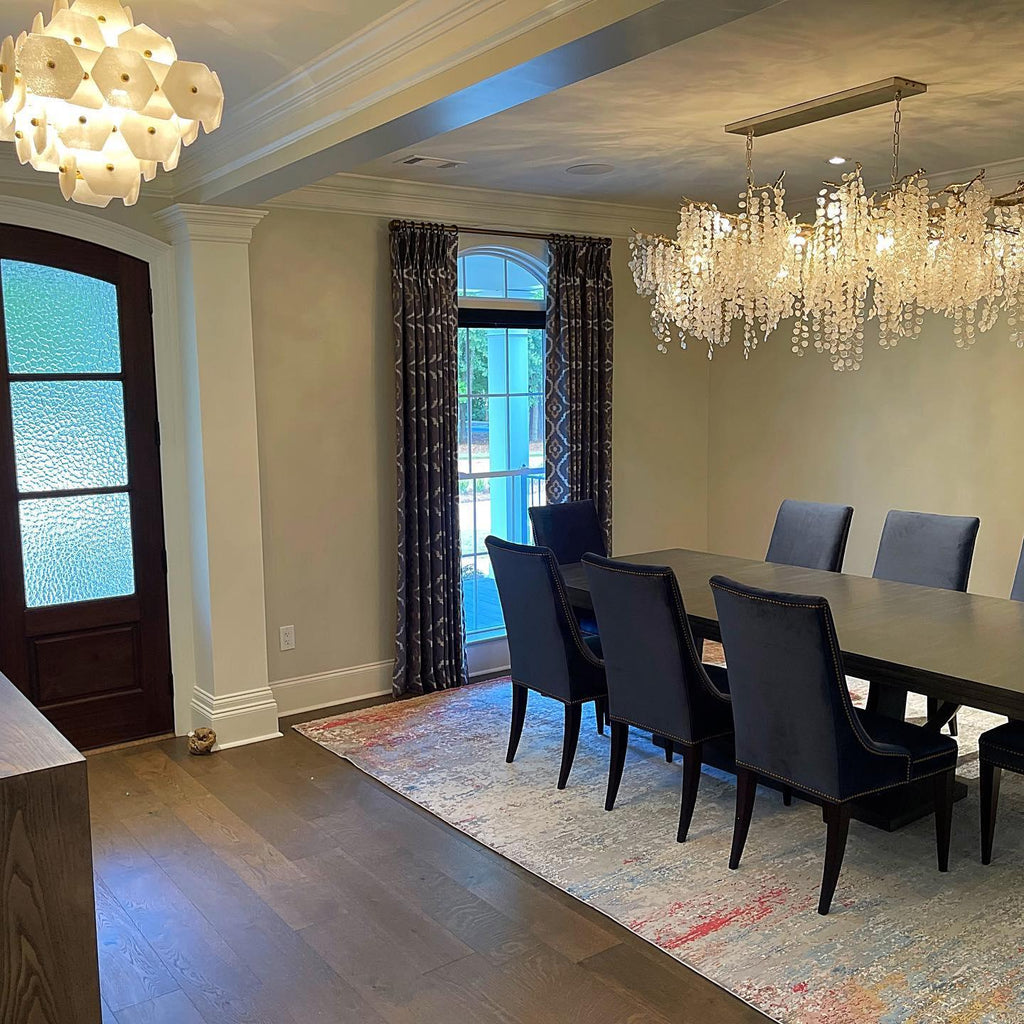 Clérachic chandelier hung in a dining room with a patterned rug, navy velvet chairs and tall windows with dark curtains.