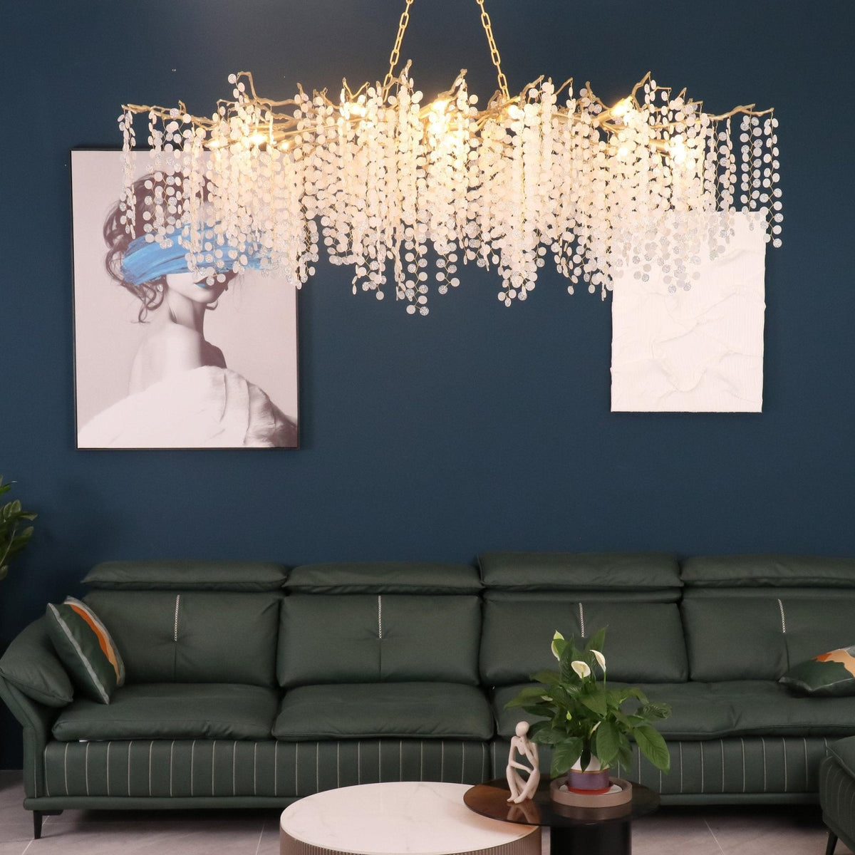 Lit Clérachic chandelier above a green leather sofa, against a teal wall with a framed portrait and a white art panel.