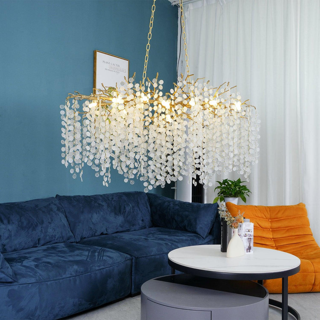 Lit Clérachic chandelier with a brass frame and warm bulbs over a navy sofa, a round coffee table and a white curtain.
