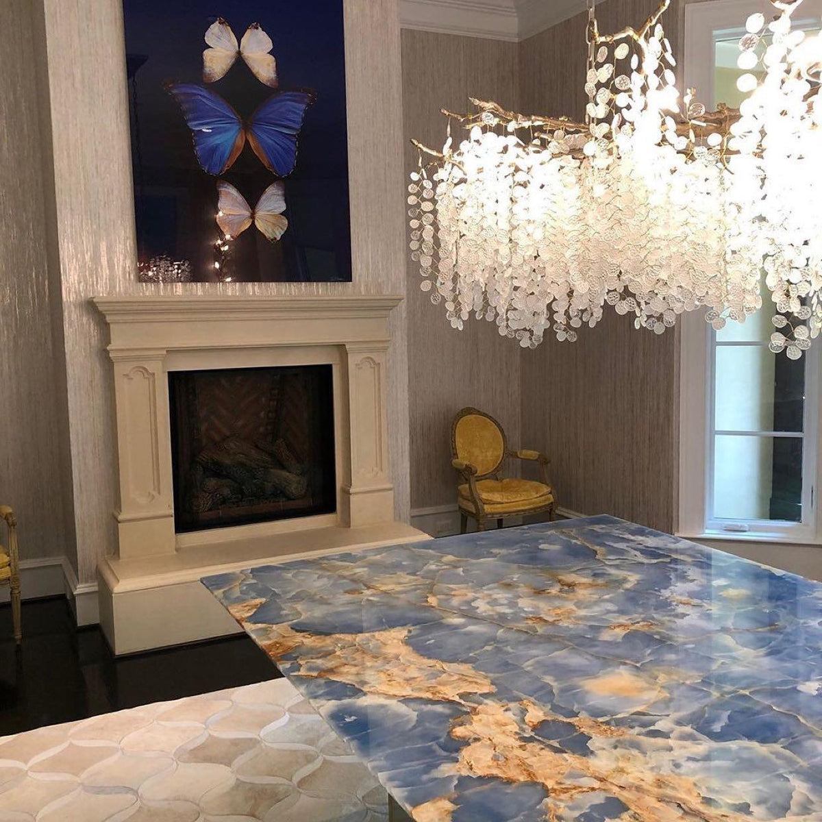 Clérachic chandelier hung beside a cream stone fireplace, with a blue butterfly artwork and a blue onyx-effect table.