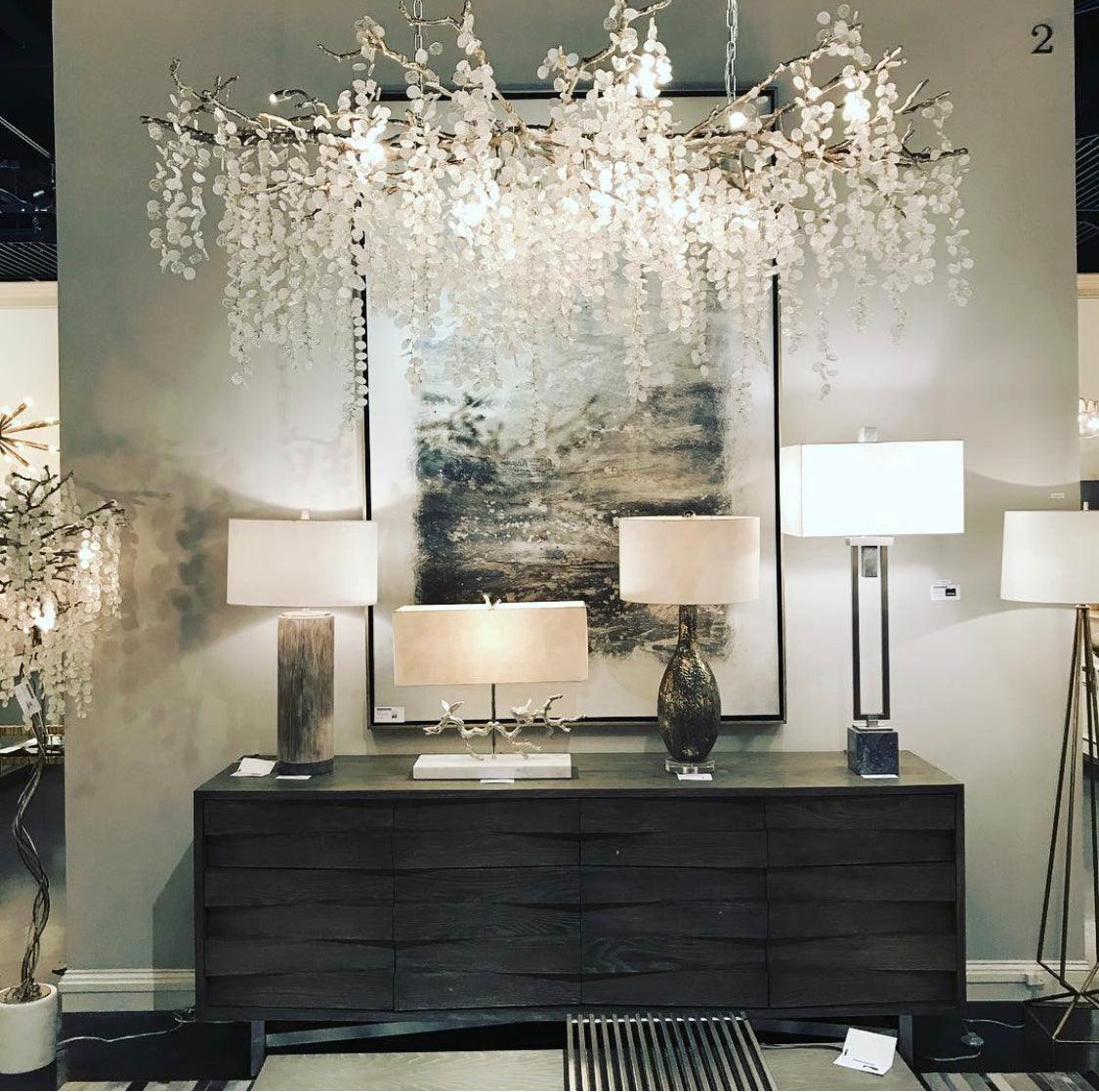 Clérachic chandelier in a showroom, lit above a dark wavy-front sideboard with table lamps and a large abstract painting.