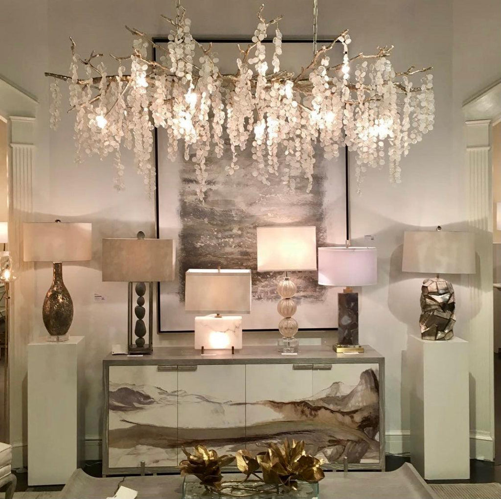 Lit Clérachic chandelier above a white sideboard painted with a landscape, with table lamps on white plinths beside it.