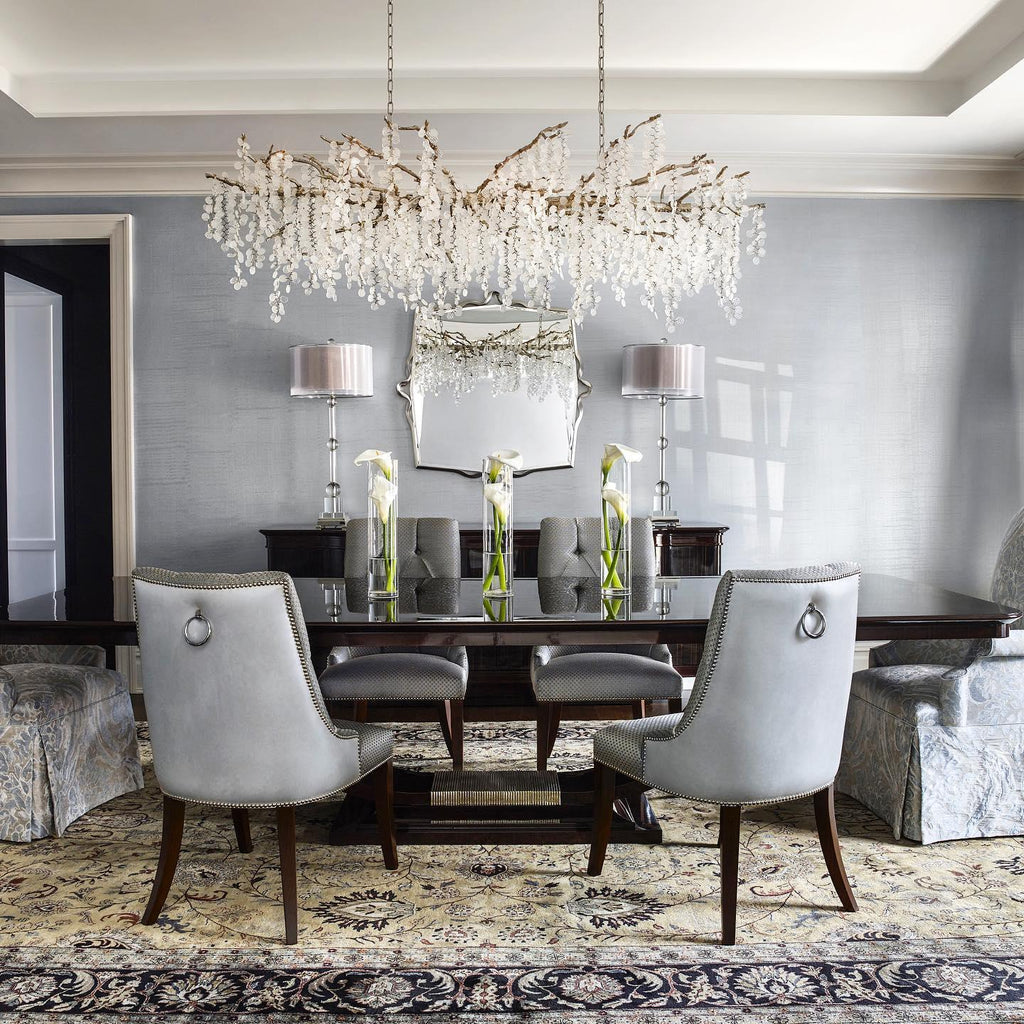 Clérachic chandelier over a dark dining table in a grey room, with pale grey upholstered chairs, a patterned rug and tall white flowers.