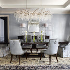 Clérachic chandelier over a dark dining table in a grey room, with pale grey upholstered chairs, a patterned rug and tall white flowers.
