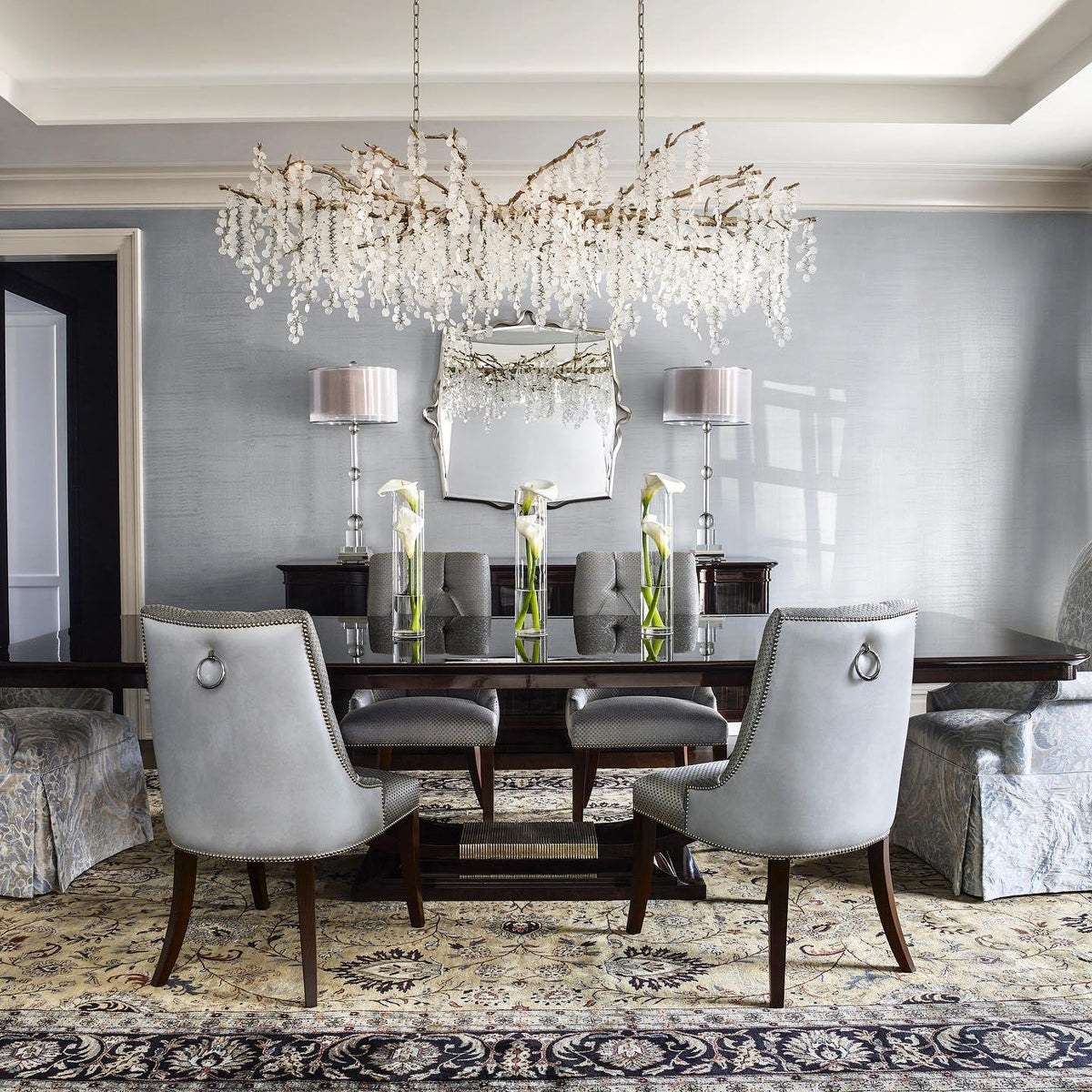 Clérachic chandelier over a dark dining table in a grey room, with pale grey upholstered chairs, a patterned rug and tall white flowers.