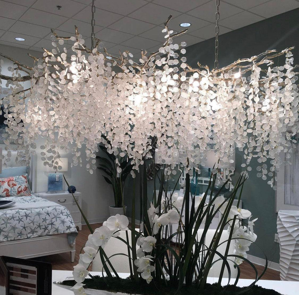 Low view of the Clérachic chandelier in a showroom, with a bedroom set and tall white orchids beneath the crystal discs.