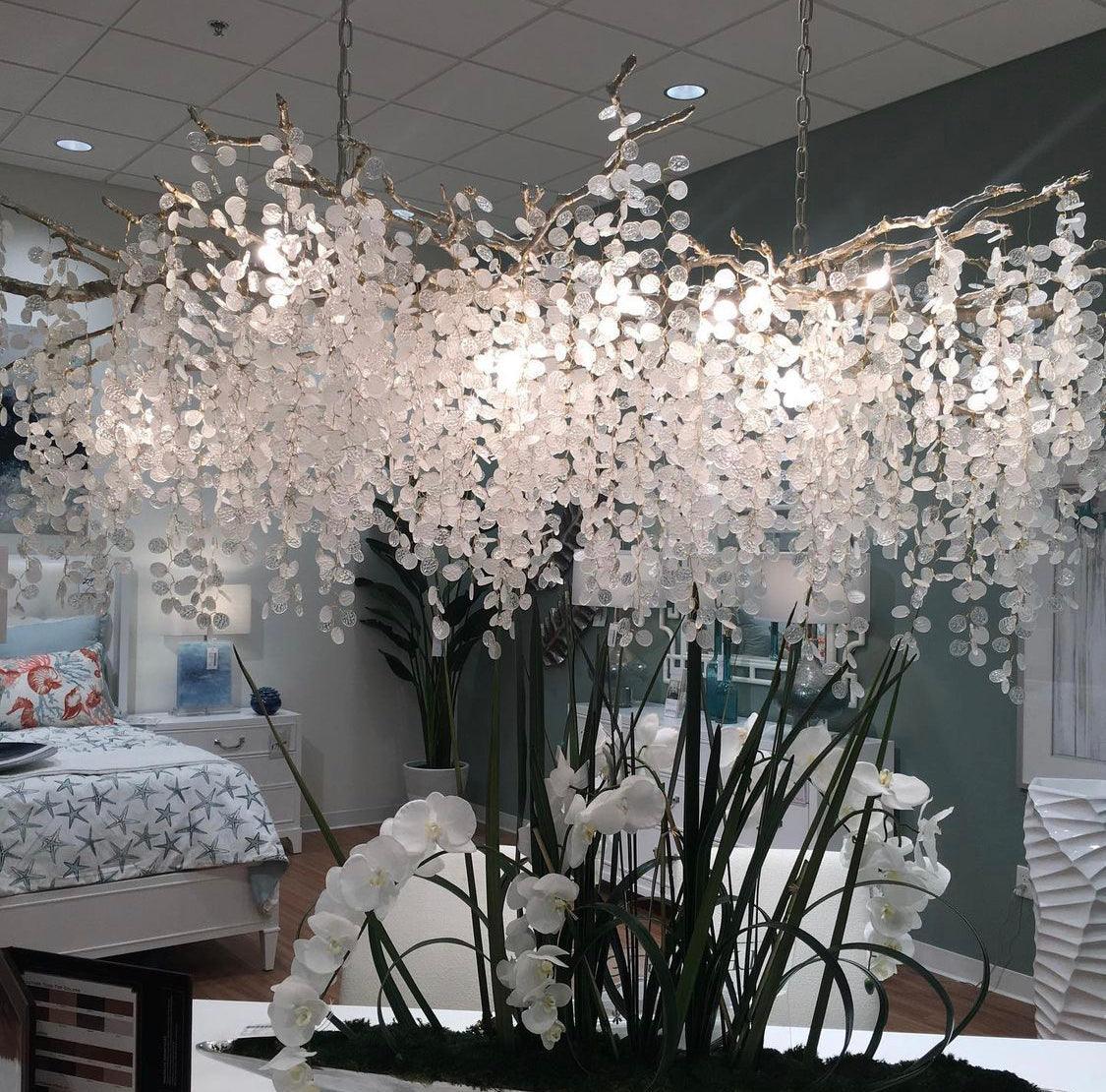 Low view of the Clérachic chandelier in a showroom, with a bedroom set and tall white orchids beneath the crystal discs.