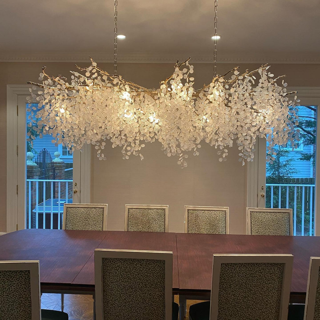 Lit Clérachic chandelier over a dining table with woven-fabric chairs, in front of two balcony doors and a white rail.