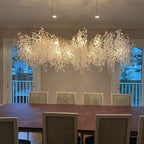 Lit Clérachic chandelier over a dining table with woven-fabric chairs, in front of two balcony doors and a white rail.
