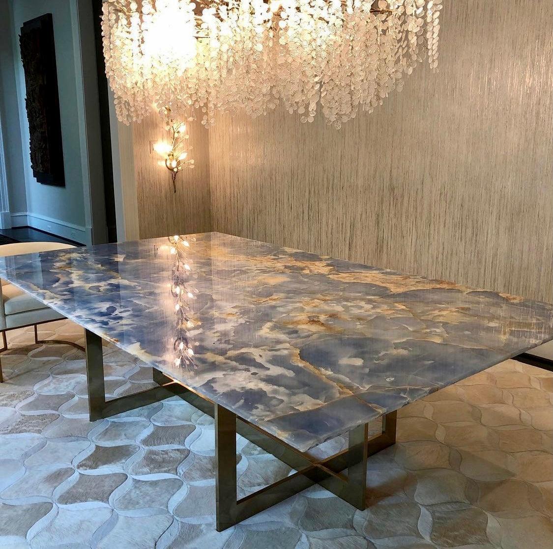 The Clérachic chandelier above a blue and gold stone table on brass legs, with a textured wall and a wall light behind.
