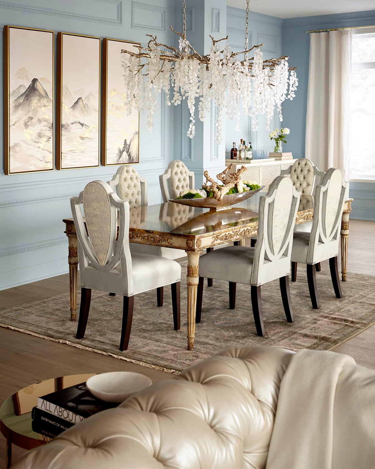 Lit Clérachic chandelier in a dining room with pale blue panelled walls, framed landscape prints and a carved gilt table.