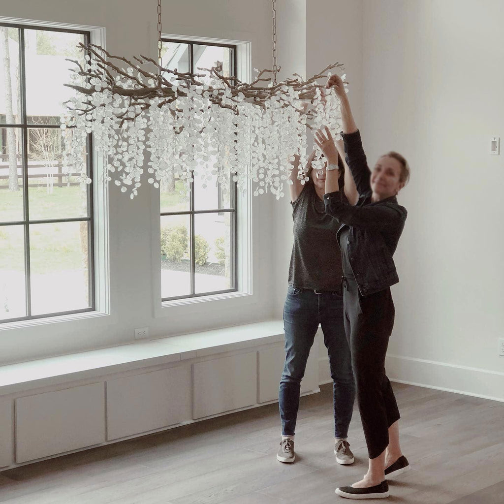 Two people reaching up to adjust the Clérachic chandelier as it hangs in an empty room with tall windows and a window seat.