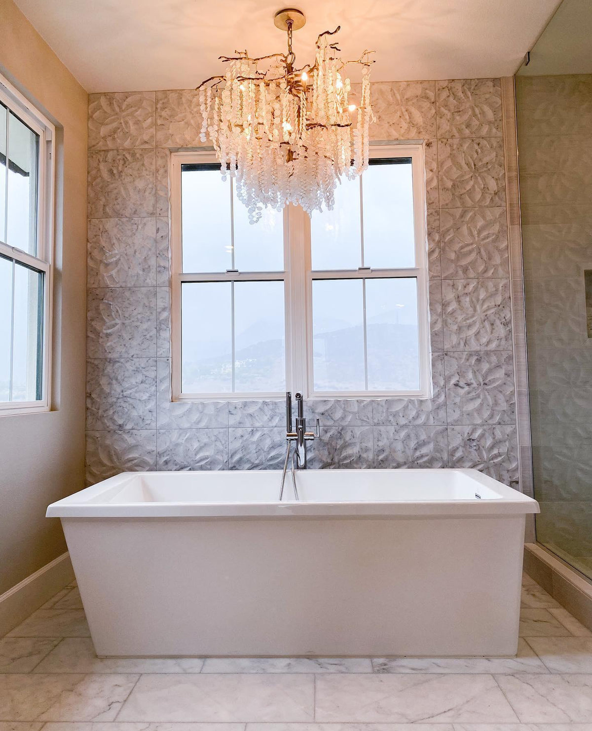 A Clérachic chandelier hung above a freestanding bath, with a window onto a hillside and a patterned tile wall.