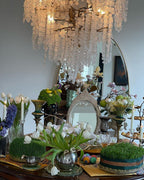 Clérachic chandelier hung above a table dressed with tulips, candles, trays of fresh grass and mirrors, with an arched mirror behind.