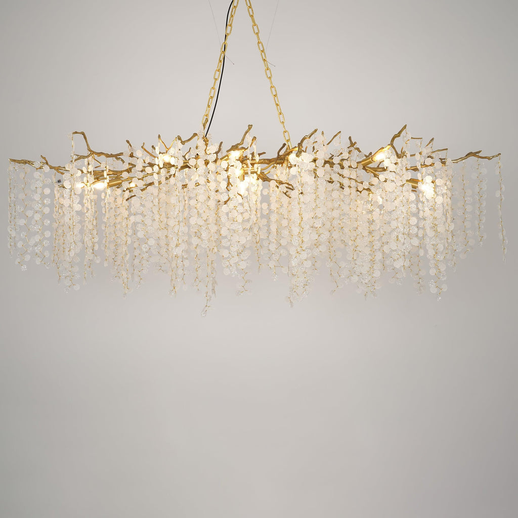 Lit Clérachic chandelier on a warm grey backdrop, with brass branches, glowing bulbs and long strands of crystal discs.