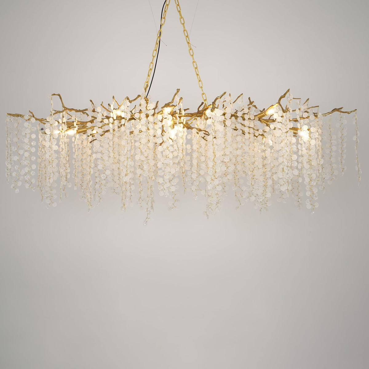 Lit Clérachic chandelier on a warm grey backdrop, with brass branches, glowing bulbs and long strands of crystal discs.