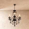 Clérachic chandelier in black glass hangs from a fine chain, three candle bulbs lit against a cream plaster wall.