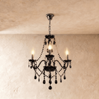 Clérachic chandelier in black glass hangs from a fine chain, three candle bulbs lit against a cream plaster wall.