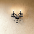 Clérachic wall light in black glass, two candle arms lit against a warm plaster wall, matching crystal drops below.