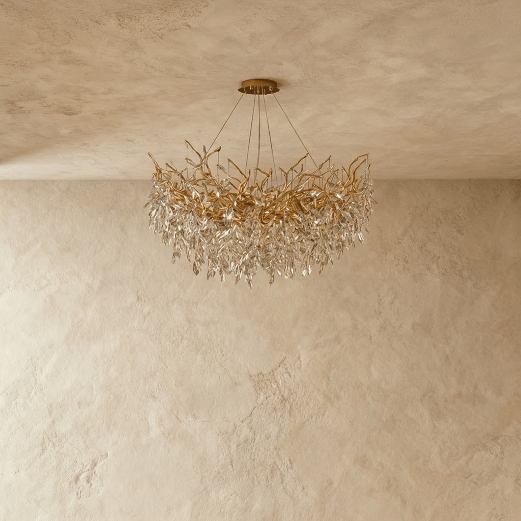 Clérachic gold chandelier with tangled branch arms and faceted crystal leaves, unlit and shown against a plaster wall.