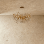 Clérachic gold chandelier with tangled branch arms and faceted crystal leaves, unlit and shown against a plaster wall.