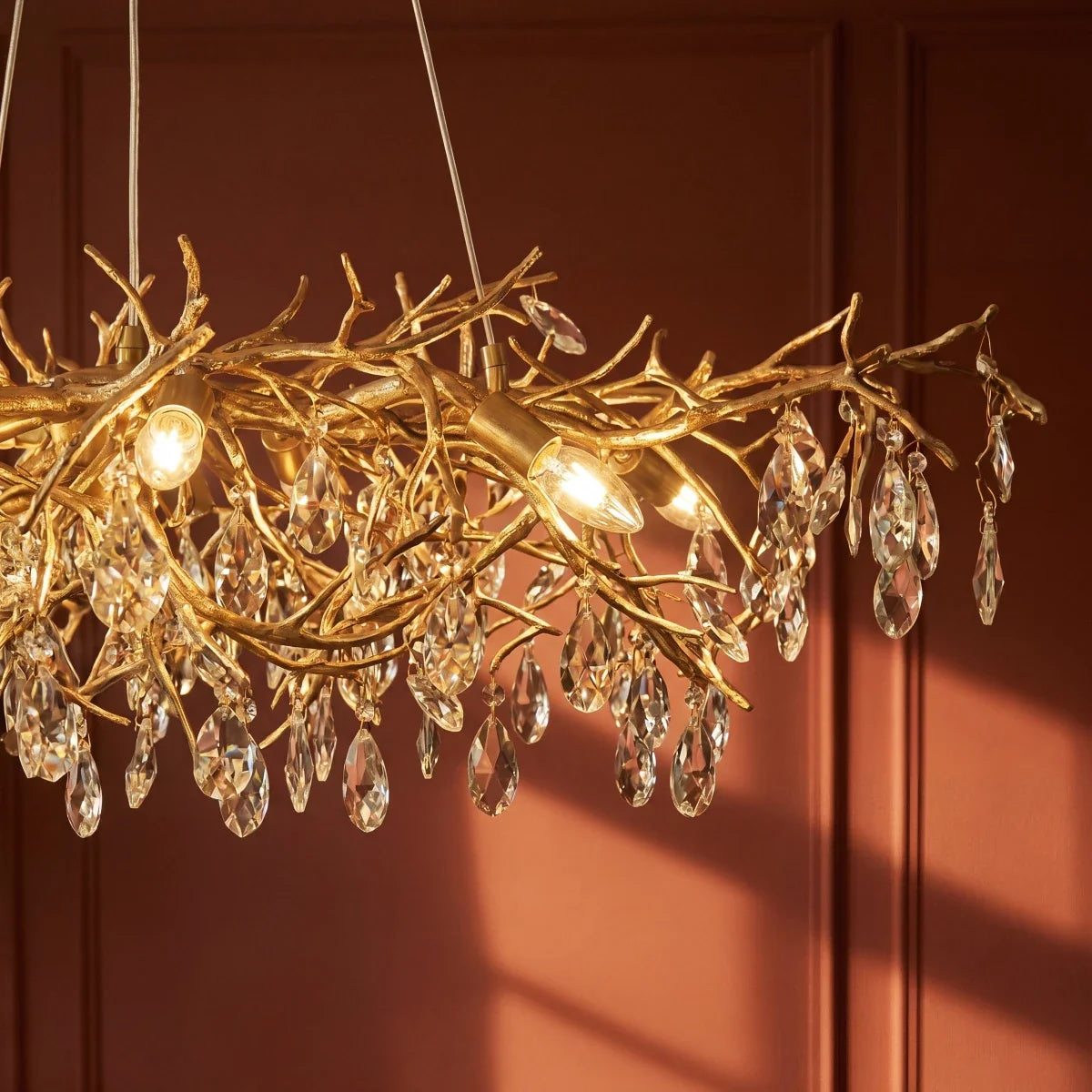 Clérachic chandelier detail showing gold branch arms hung with faceted crystal drops against a terracotta wall.