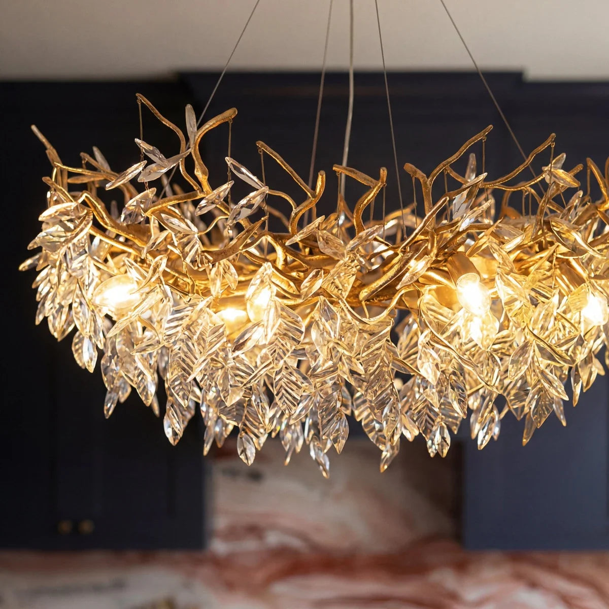 Clérachic chandelier detail, gold branches and crystal leaves glowing warm against dark navy kitchen cabinets.