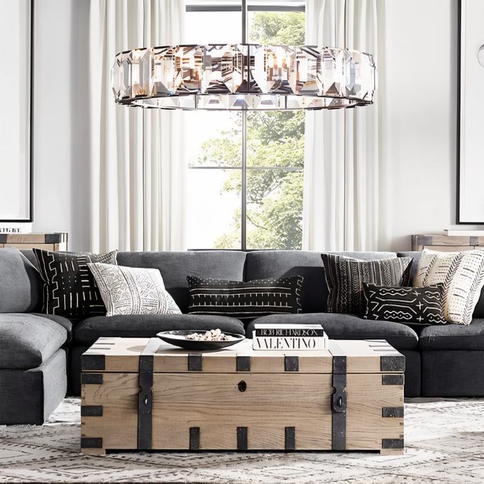 Clérachic wide ring chandelier of faceted crystal hanging above a charcoal sofa and wooden trunk table by a tall window.